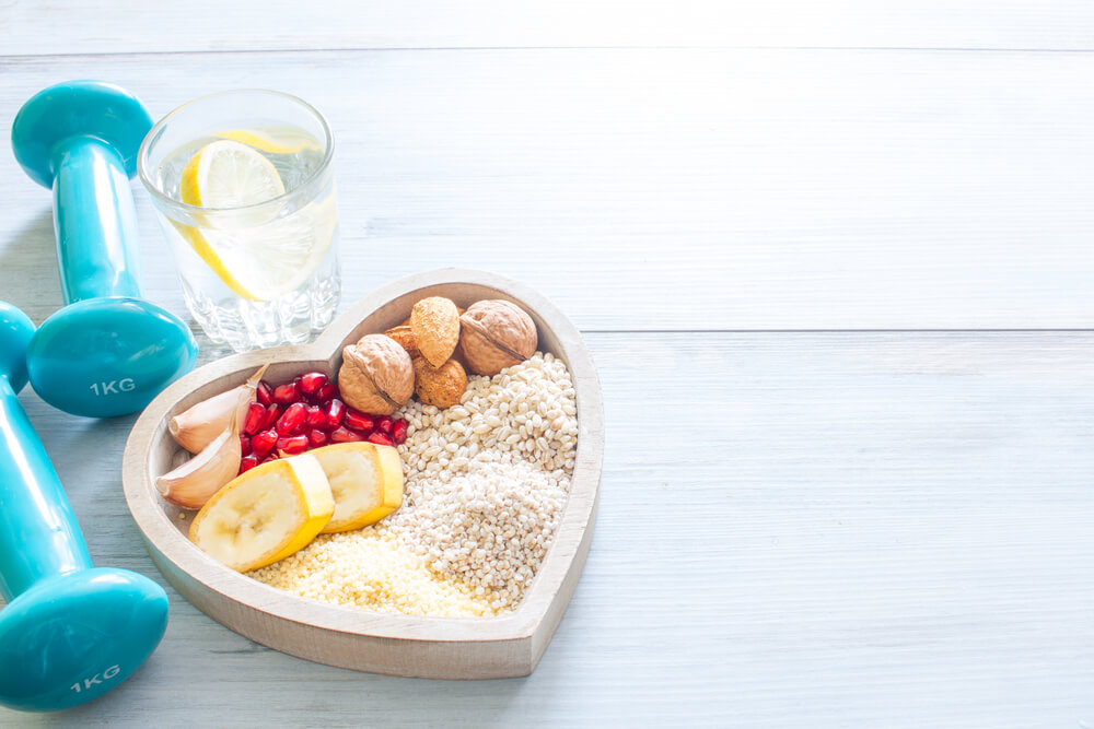 Plato en forma de corazón con alimentos saludables junto a unas mancuernas y un vaso de agua con limón