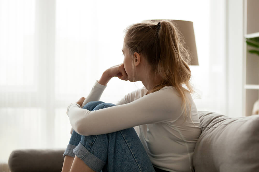 Mujer joven sentada en el sofá con las rodillas dobladas y mirando a la ventana