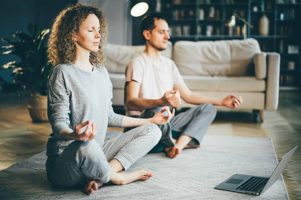 Pareja haciendo meditación en la alfombra de la sala de estar frente a una computadora portátil.