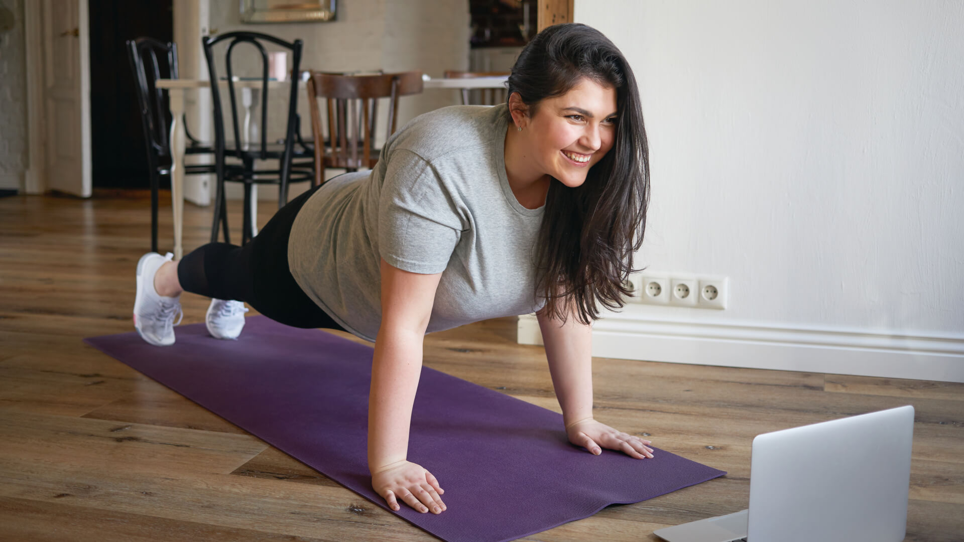 Mujer joven con sobrepeso haciendo yoga en casa frente a una computadora.